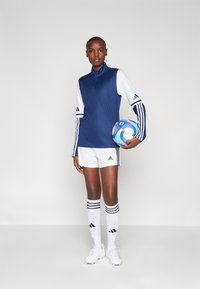Marineblå og hvid atletisk top med lynlås, hvide shorts og stribede knæstrømper. Model holder en multikolor fodbold.