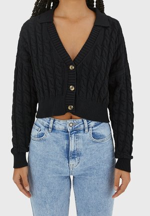 Personne portant un cardigan court en maille torsadée noire avec des boutons et un jean taille haute bleu clair sur un fond uni.