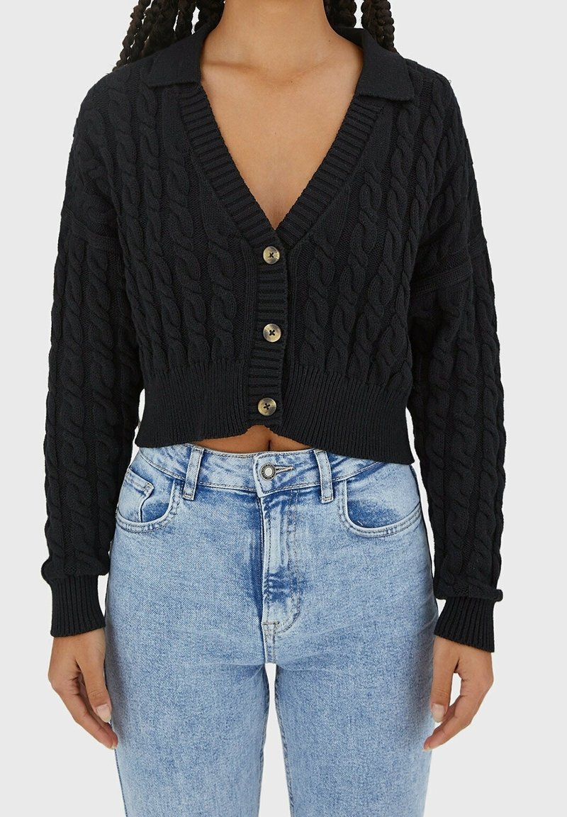Personne portant un cardigan court en maille torsadée noire avec des boutons et un jean taille haute bleu clair sur un fond uni.