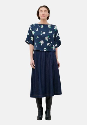 Femme portant un haut à fleurs bleu marine à manches courtes, une jupe midi bleu foncé et des bottes noires montantes, debout devant un fond uni.