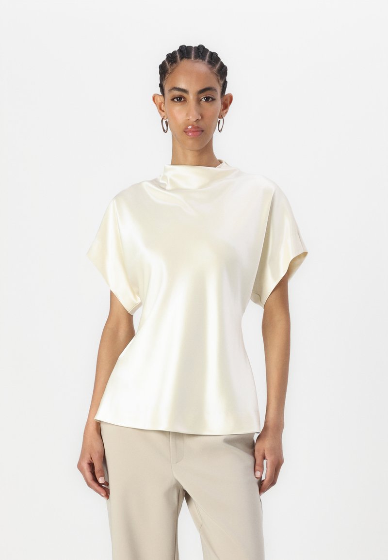 Krem satar topp med høy hals og korte, brede ermer. Glatt tekstur, avslappet passform og elegant fall. Kombinert med lyse beige bukser.