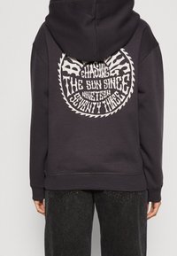 Osoba oblečená v černé mikině s kapucí, na zádech s bílým kruhovým grafickým textem "Honíme slunce od devatenáct set sedmdesáti tří".