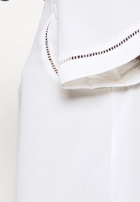 Blouse blanche avec un col texturé présentant des découpes décoratives et un design fluide. Fabriquée en tissu léger avec un tomber doux.