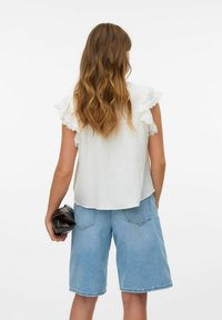 Vit strukturerad topp med spetsbeskurnade ärmar, i kombination med ljusblå denimshorts. Individuen håller en svart clutch i vänster hand.