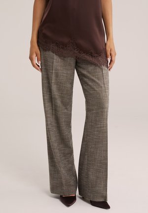 Femme portant un pantalon gris à carreaux à jambes larges, un haut marron foncé orné de dentelle et des chaussures pointues marron foncé, debout devant un fond uni.