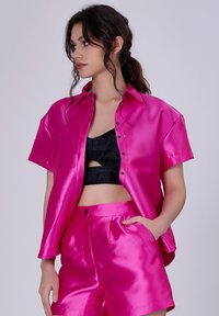 Chemise satin fuchsia à manches courtes avec un col pointu, associée à un short assorti. Débardeur noir en dessous. Présente une texture brillante et des détails de boutons.