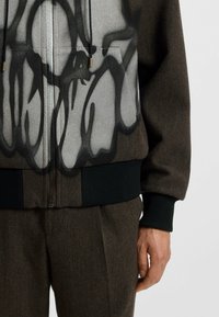 Kapturowa kurtka z szaro-czarnym abstrakcyjnym wzorem, wykonana z teksturowanej tkaniny, z prążkowanymi mankietami i dołem w ciemnozielonym kolorze.