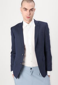 Navy textured blazer with two buttons, tailored fit, worn over a white shirt. Blue trousers with a flat front and visible button closure.