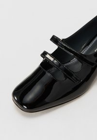 Black patent leather shoe with a squared toe, featuring two adjustable straps across the instep and a smooth inner lining.