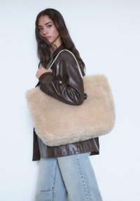 Béžová kabelka z umelého kožucha s obdĺžnikovým tvarom, mäkkou textúrou a svetlými koženými popruhmi. Nošená ležérne cez plece.
