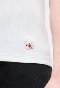 Κλείσιμο ενός ατόμου που φοράει λευκό ριμπάν πλεκτό τοπ με ένα μικρό κόκκινο και μαύρο λογότυπο Calvin Klein κοντά στο τελείωμα, σε συνδυασμό με μαύρο παντελόνι.