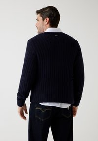 Uomo in un pullover a coste blu navale sopra una camicia bianca e jeans scuri, mostrato di spalle su uno sfondo chiaro e semplice.