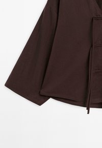 Haut à manches longues marron foncé avec manches larges et fermeture nouée à l'avant, présenté sur fond blanc.