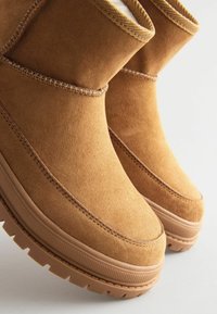 Brune ruskindsankelstøvler med en struktureret overflade, gummisåler og minimal syning. Har et let hævet, rundt design.