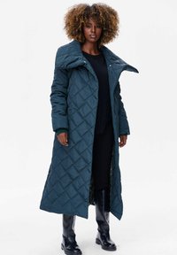 Lange, gewatteerde jas in diep teal met een grote kraag, voorzien van een gladde textuur en zwarte binnenvoering. Draagt over een donkere trui en een jurk.