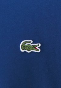 Tissu en coton bleu marine avec un petit logo crocodile vert brodé et un accent rouge, positionné de manière visible sur le vêtement.