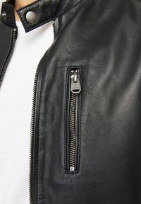 Jachetă din piele neagră cu un buzunar cu fermoar pe partea stângă, accesorii din metal argintiu și închidere cu capse la guler. Textură netedă.