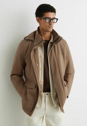 Muž nosiaci čierne okuliare, hnedú vrstvu bundy cez béžové pletené tričko a krémové nohavice, stojí pri bielej stene, ruky v kapsách.