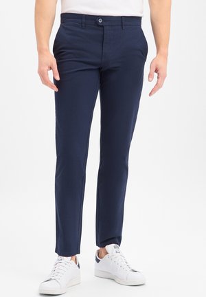 Man draagt slim-fit marineblauwe broek en witte sneakers en staat tegen een witte achtergrond.