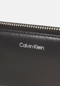 Portafoglio in pelle nera con zip argentata e logo bianco "Calvin Klein" sulla parte anteriore. Texture liscia con dettagli a grana sottile.