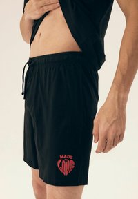 Czarne szorty wykonane z lekkiego materiału, z elastycznym paskiem w talii i sznurkiem do regulacji oraz czerwonym napisem "MADE TO LOVE" na dolnej części.