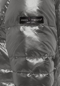 Szara kurtka puchowa z błyszczącego materiału, z pikowanym wzorem i czarną metką z logo "FRIEDA FREDDIES NEW YORK".