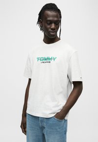 Ανοιχτό γκρι βαμβακερό μπλουζάκι με τύπωμα λογοτύπου σε πράσινο και μαύρο, με χαλαρή εφαρμογή και κοντά μανίκια. Τζιν παντελόνι εμφανές στο κάτω μέρος.