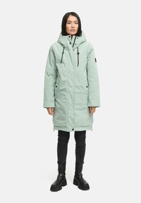 Hellgrüne, kapuzenbehangene, lange Parkajacke mit schwarzen Akzenten, Frontreißverschluss und Druckknopfverschluss; getragen über dunklen Jeans und schwarzen Stiefeln.