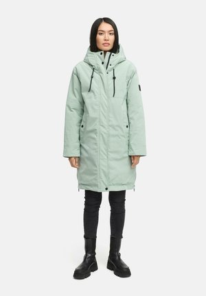 Lichtgroene, met capuchon, lange parka-jas met zwarte accenten, rits aan de voorkant en drukknoopsluiting; gedragen over donkere jeans en zwarte laarzen.