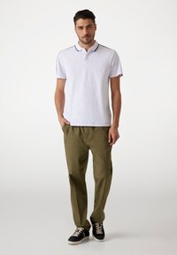 Camisa polo blanca con cuello y ribetes negros, combinada con pantalones verde oliva. El modelo lleva zapatillas negras con suela blanca.