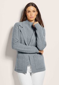 Grijze gebreide cardigan met een sjalkraag, geribbelde mouwen en zijzakken. Zachte textuur en open ontwerp, gelaagd over een turtleneck.