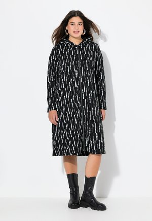 Manteau à capuche zippé noir avec un motif abstrait blanc, à manches longues, atteignant mi-mollet, associé à des bottes noires jusqu'au genou.