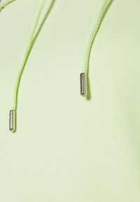 Felpa verde lime in un tessuto morbido, con lacci intrecciati e una finitura metallica sulle punte. Primo piano sui dettagli del materiale e del design.