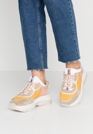 Person wearing yellow, beige, and pink chunky sneakers with white soles and laces, paired with cropped blue denim jeans.