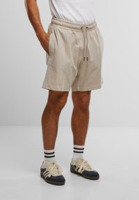 Beige Baumwollshorts mit einem elastischen Bund und Kordelzug. Sie verfügen über seitliche Taschen und einen entspannten Schnitt. Getragen mit marineblauen und cremefarbenen Sneakern.