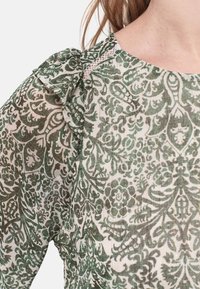 Blusa verde e bianca a motivo, con dettagli arricciati sulle spalle, motivi floreali e un tessuto leggero e testurizzato.