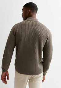 Brown ribbed knit sweater with a high collar, long sleeves, and a relaxed fit, paired with light-colored pants.