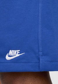 Pantaloni corti Nike blu con una texture liscia, dotati di un logo ricamato bianco vicino all'orlo e cuciture a contrasto lungo le cuciture.