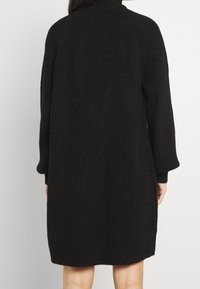 Robe pull noire en maille oversize avec un col montant, des manches longues et un tissu texturé. Elle présente une coupe décontractée et des poignets côtes.
