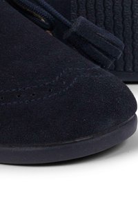 Zapato de ante azul marino con costuras visibles, punta redonda, suela plana de goma y cordones en contraste con un adorno de borlas decorativas.
