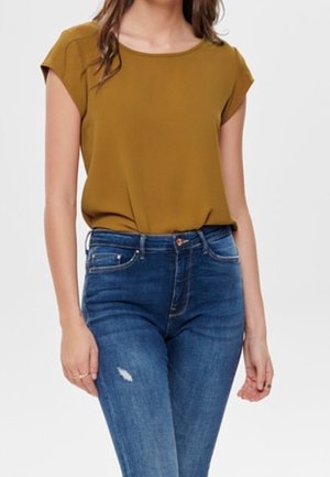 Vrouw draagt een mosterdkleurige blouse met korte mouwen, ingestopt in een hoog opgesneden blauwe spijkerbroek met lichte slijtage op het bovenbeen.