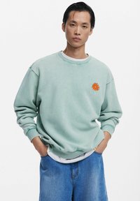 Sweat-shirt vert clair avec un col rond, arborant un logo de fleur orange sur la poitrine gauche. Porté sur une chemise blanche et associé à un short en jean bleu.