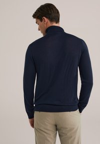 Hombre visto desde atrás, vistiendo un suéter de cuello alto azul marino y pantalones beige, contra un fondo neutro y simple.