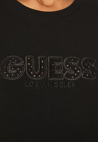 Camiseta de algodón negra con "GUESS" en letras gruesas y texturizadas, adornada con tachuelas superpuestas y "LOS ÁNGELES" en una fuente más pequeña debajo.