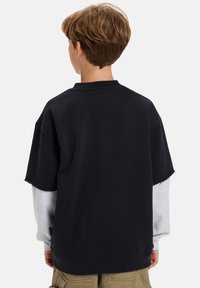 Giovane con capelli castano chiaro corti, che indossa una camicia nera a maniche corte a strati sopra una camicia grigia a maniche lunghe, di spalle.