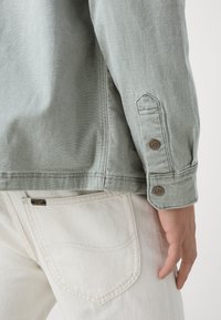 Primer plano de la mano de una persona descansando a su lado, vistiendo una chaqueta verde salvia y pantalones beige claros con etiquetas de la marca Lee en el puño y el bolsillo.