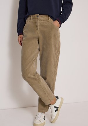 Personne portant un pantalon en velours côtelé beige, un pull bleu marine et des baskets blanches avec des détails noirs, debout avec une main dans la poche.