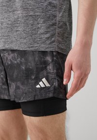 T-shirt de sport gris en tissu texturé, associé à un short noir avec un motif marbré et un logo Adidas blanc.