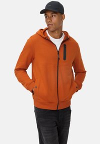 Orange zip hoodie med en sort lynlås lomme, ribbede manchetter og nederste kant. Parret med mørke jeans. Stofoverfladen fremstår glat og let struktureret.