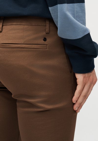 Καφέ παντελόνι ραμμένο στα μέτρα με πίσω τσέπη welt και μαύρο κουμπί, συνδυασμένο με μπλε και μαύρο μακρυμάνικο μπλουζάκι, το χέρι ακουμπάει στον μηρό.